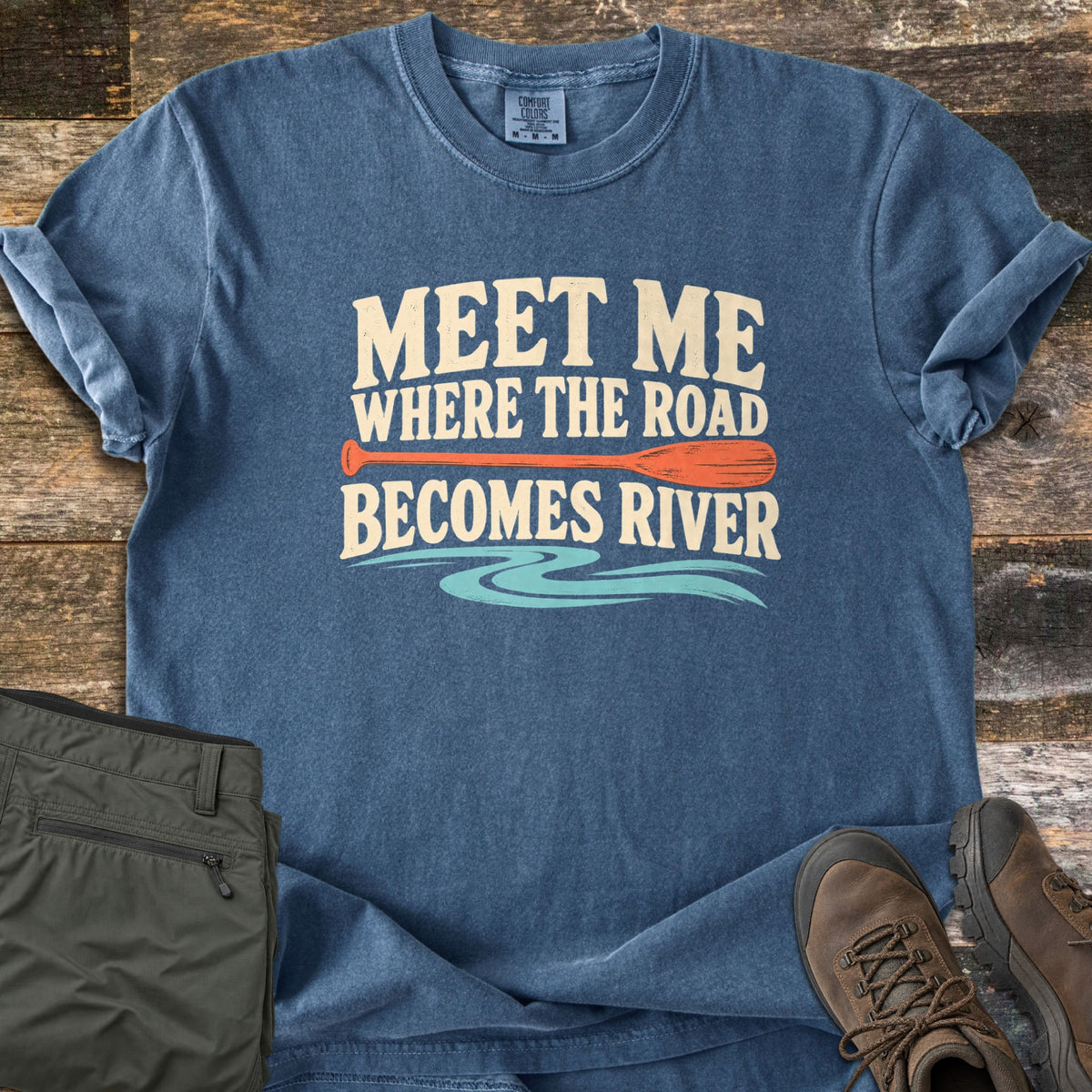 Meet Me Where the Road Becomes River Comfort Colors T-shirt in Denim, laid flat. Cream text reads Meet Me Where the Road Becomes River, with an orange canoe paddle between the lines and turquoise water swirls below. Sleeves are rolled on wood planks beside olive hiking pants and brown boots.
