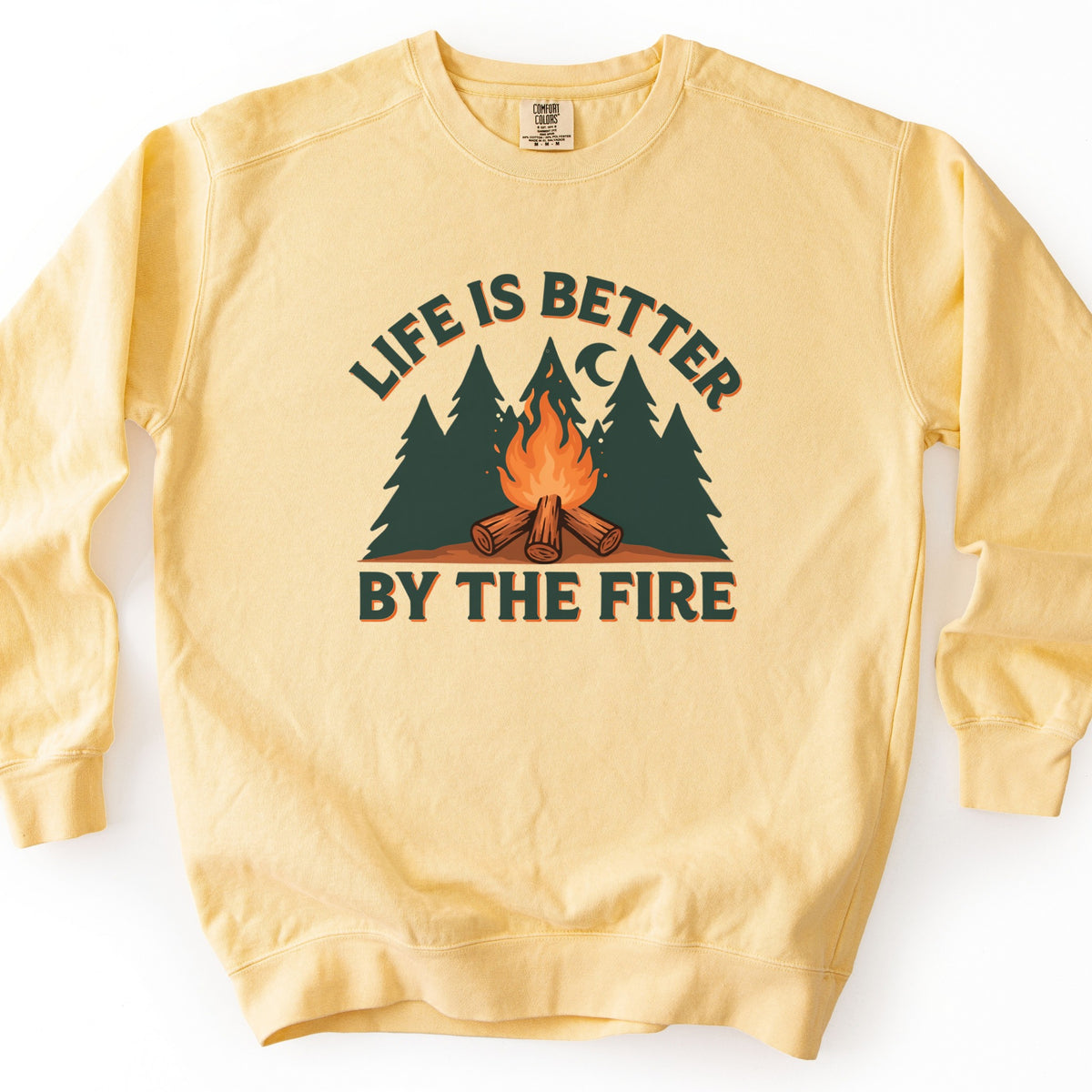Life Is Better By The Fire Comfort Colors crewneck sweatshirt in Butter, laid flat. Dark green letters with an orange shadow arch Life Is Better over dark green pine trees, a crescent moon and a campfire of crossed logs, with By The Fire below. It lies on a white surface.