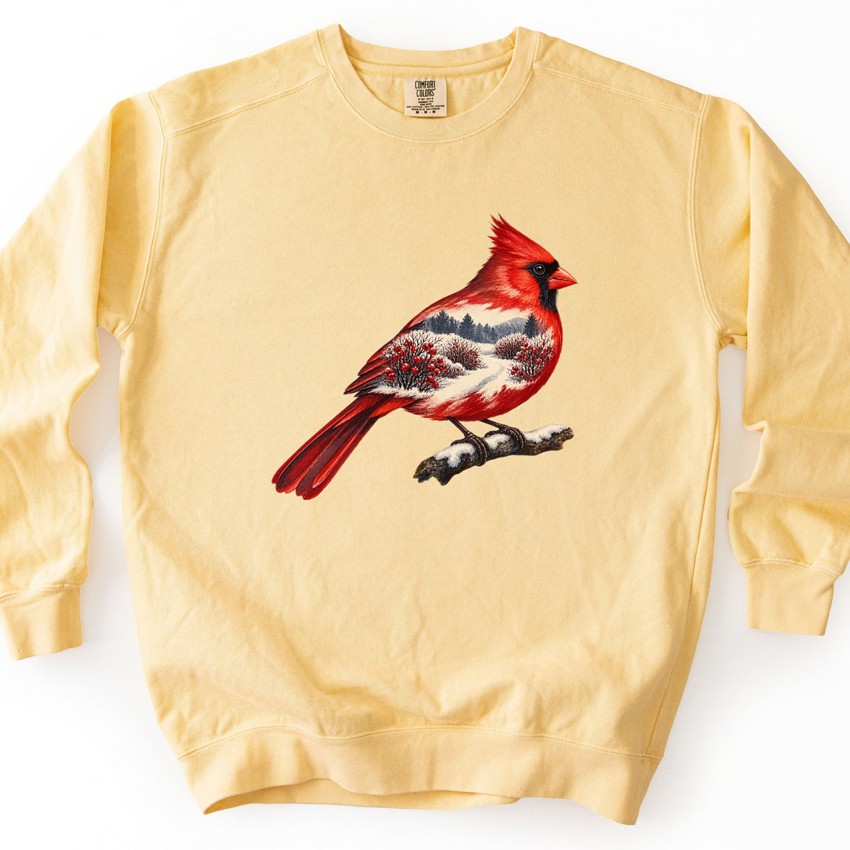 Winter Cardinal Comfort Colors crewneck sweatshirt in Butter, laid flat. A painted red cardinal perched on a snowy branch, with a winter scene of snow, pine trees and red berry bushes inside its body. No words are printed. Shown on a white background.