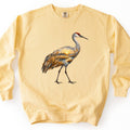 Crane Marsh Sunrise Comfort Colors crewneck sweatshirt in Butter, laid flat. A sandhill crane with a red crown walks in profile, its body filled with a marsh sunrise scene of golden reeds, a winding stream and trees under a glowing sun. No words are printed. Shown on a white background.