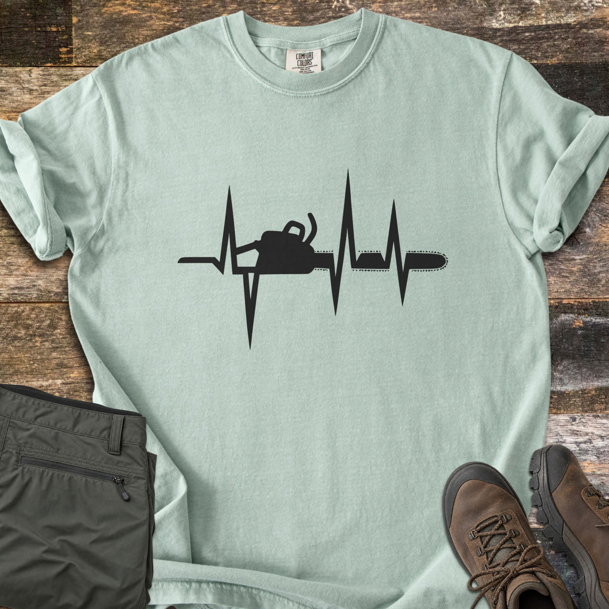 Chainsaw Heartbeat Comfort Colors T-shirt in Bay, laid flat. A black heartbeat line runs across the chest with a chainsaw silhouette built into it, the bar and chain extending to the right. Folded olive cargo shorts and brown hiking boots sit on a weathered wood floor.