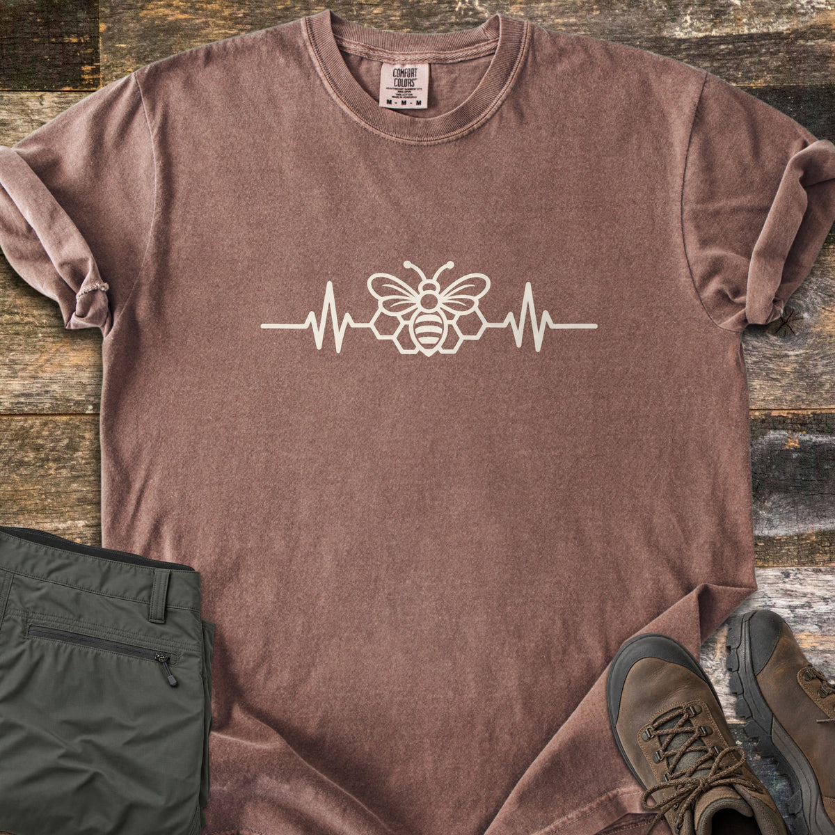 Bee Heartbeat Comfort Colors T-shirt in Espresso, laid flat. A cream line drawing of a bee over honeycomb hexagons sits in the middle of a heartbeat line across the chest. Sleeves are rolled, on weathered wood beside olive hiking pants and brown hiking boots.