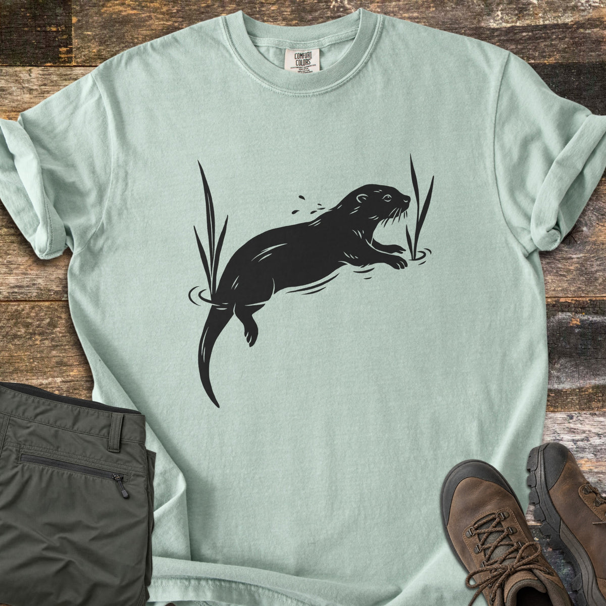 Swimming Otter Comfort Colors T-shirt in Bay, laid flat. A black print of an otter swimming between tall reeds, with ripples and splashing droplets. Sleeves are rolled; olive hiking pants and brown hiking boots sit on weathered wood planks.