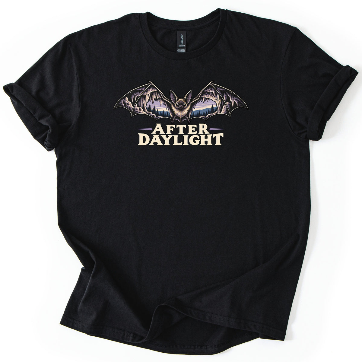 After Daylight Comfort Colors T-shirt in Black, laid flat. A fuzzy bat with outstretched wings, each wing filled with a cave scene of stalactites and a purple dusk sky over pine trees, above the words After Daylight in cream letters. Sleeves rolled, on a white background.