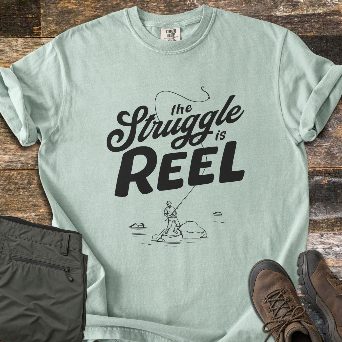 The Struggle Is Reel Comfort Colors T-shirt in Bay, laid flat. Black text reads the Struggle is REEL under an arcing fishing line with a hook, above a sketch of an angler casting from a rock in water. Folded olive pants and brown hiking boots sit on wood planks.