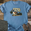 Fowl Play Comfort Colors T-shirt in Blue Jean, laid flat. A cartoon Canada goose carries a green off-road helmet in its beak past a green ATV and pine trees, under the words Fowl Play. Sleeves rolled, on weathered wood with olive pants and brown hiking boots.