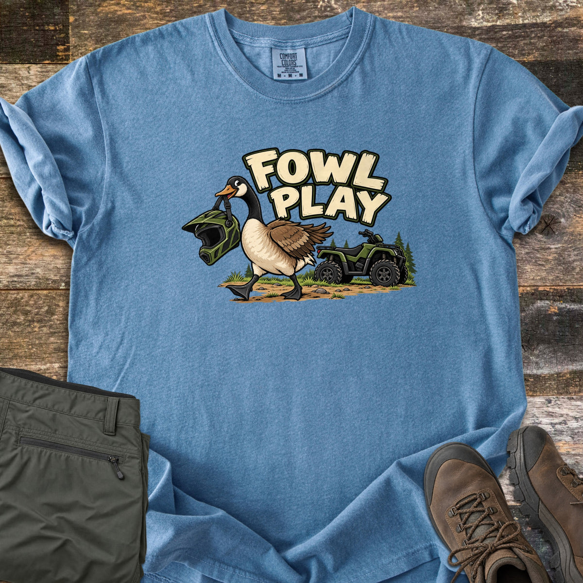 Fowl Play Comfort Colors T-shirt in Blue Jean, laid flat. A cartoon Canada goose carries a green off-road helmet in its beak past a green ATV and pine trees, under the words Fowl Play. Sleeves rolled, on weathered wood with olive pants and brown hiking boots.