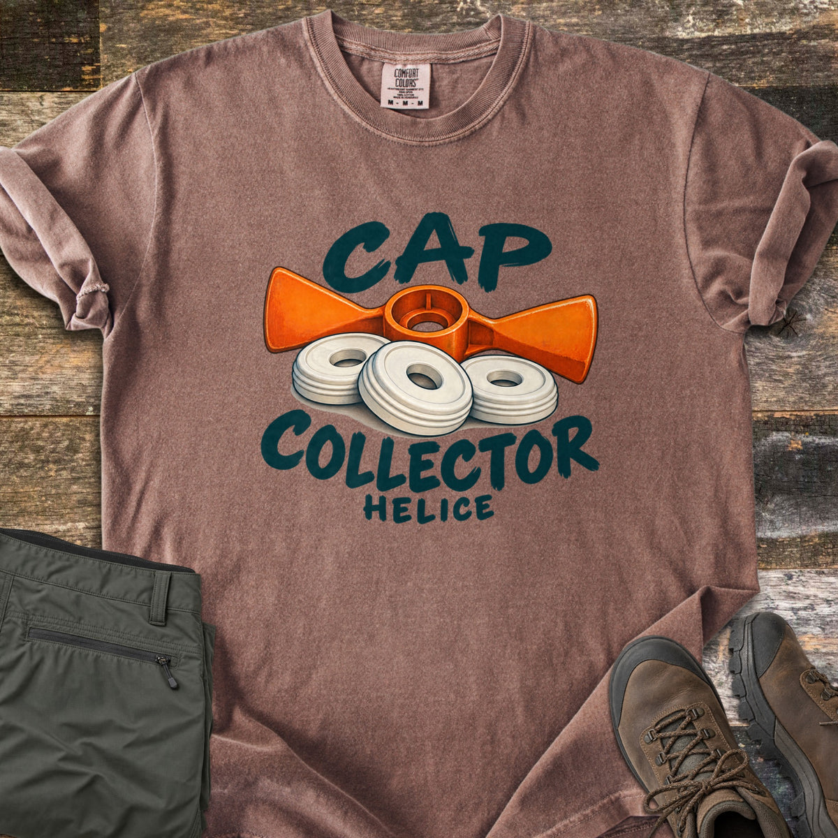 Cap Collector Comfort Colors T-shirt in Espresso, laid flat. An orange helice key and three white caps sit between the words Cap Collector, with Helice below. Sleeves are rolled; olive pants and brown hiking boots lie beside it on dark wood planks.