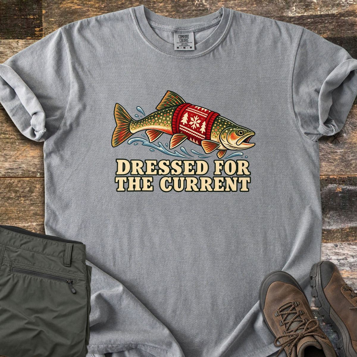Dressed for the Current Comfort Colors T-shirt in Grey, laid flat. A speckled trout wearing a red knit sweater with a white snowflake and tree pattern leaps through splashing water above the words Dressed for the Current. Olive pants and brown hiking boots lie on weathered wood.