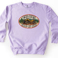 The Dirt Knows My Name Comfort Colors crewneck sweatshirt in Orchid, laid flat. An oval patch-style design shows a mud-covered yellow ATV on a dirt trail with pine trees and mountains behind it, framed by the words The Dirt Knows My Name. The sweatshirt lies on a white background.