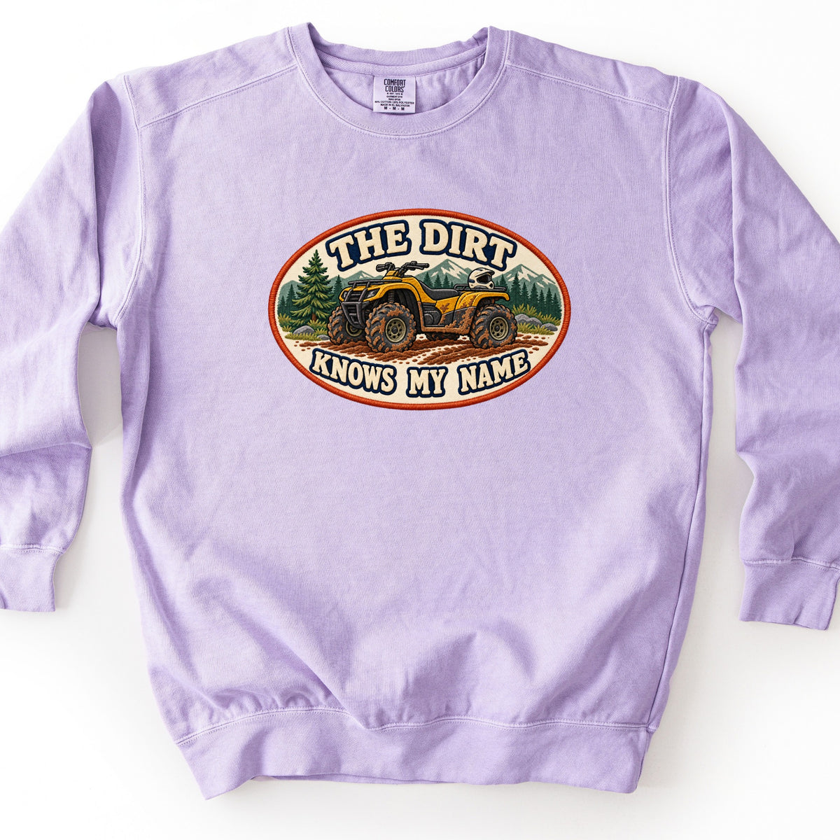 The Dirt Knows My Name Comfort Colors crewneck sweatshirt in Orchid, laid flat. An oval patch-style design shows a mud-covered yellow ATV on a dirt trail with pine trees and mountains behind it, framed by the words The Dirt Knows My Name. The sweatshirt lies on a white background.