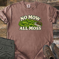 No Mow All Moss Comfort Colors T-shirt in Espresso, laid flat. Mounds of green moss with a stone path and an orange push reel mower sit between the cream words No Mow All Moss. The shirt has rolled sleeves on weathered wood beside folded olive pants and brown hiking boots.