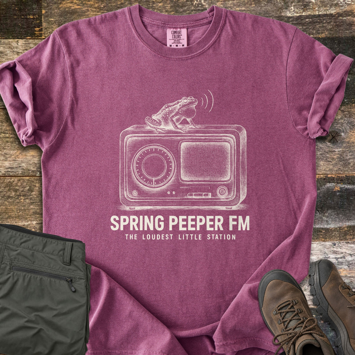 Spring Peeper FM Comfort Colors T-shirt in Berry, laid flat. A sketch of a frog with puffed throat sits on a vintage radio, sound waves above it, over the words Spring Peeper FM, The Loudest Little Station. Sleeves are rolled on wood planks beside green pants and hiking boots.