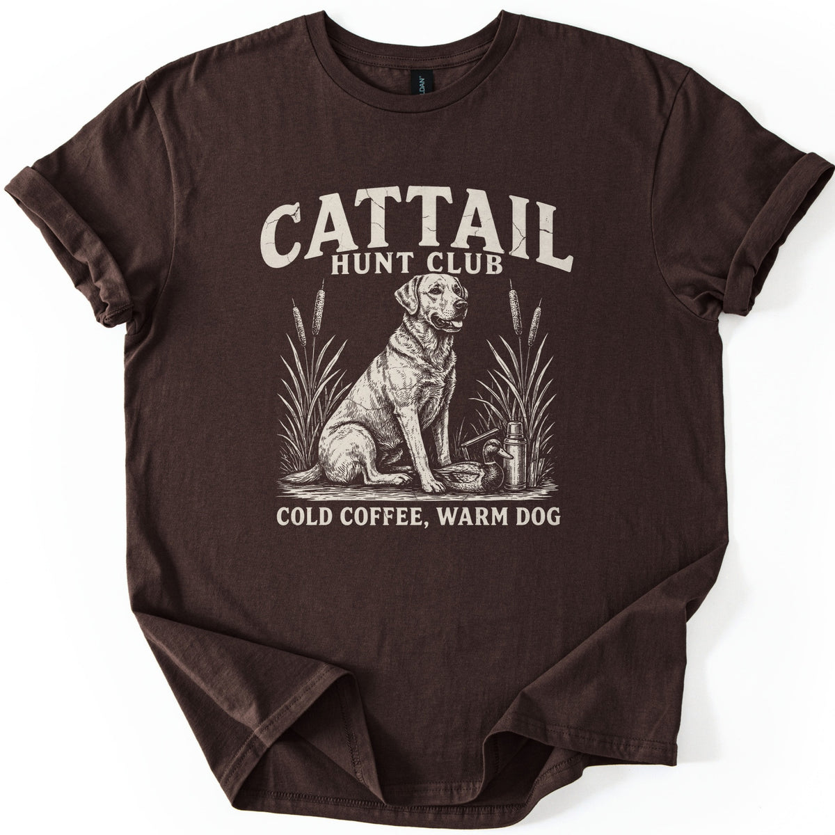 Cattail Hunt Club Comfort Colors T-shirt in Dark Chocolate, laid flat. Cream print of a Labrador retriever sitting among cattails beside a duck decoy and thermos, under the words Cattail Hunt Club and above Cold Coffee, Warm Dog. Sleeves cuffed, on a white background.