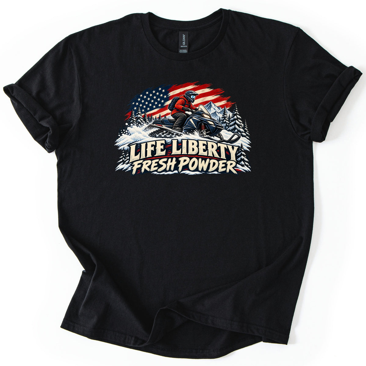 Life Liberty Fresh Powder Comfort Colors T-shirt in Black, laid flat. A rider in a red jacket and helmet kicks up snow on a snowmobile in front of an American flag, pine trees and a mountain, above the words Life Liberty Fresh Powder. Sleeves rolled, on a white background.