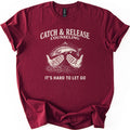 Catch & Release Counseling Comfort Colors T-shirt in Maroon, laid flat. Cream print reads CATCH & RELEASE COUNSELING above an etched trout leaping from two open hands over rippling water, with IT'S HARD TO LET GO below. Sleeves are cuffed on a white background.
