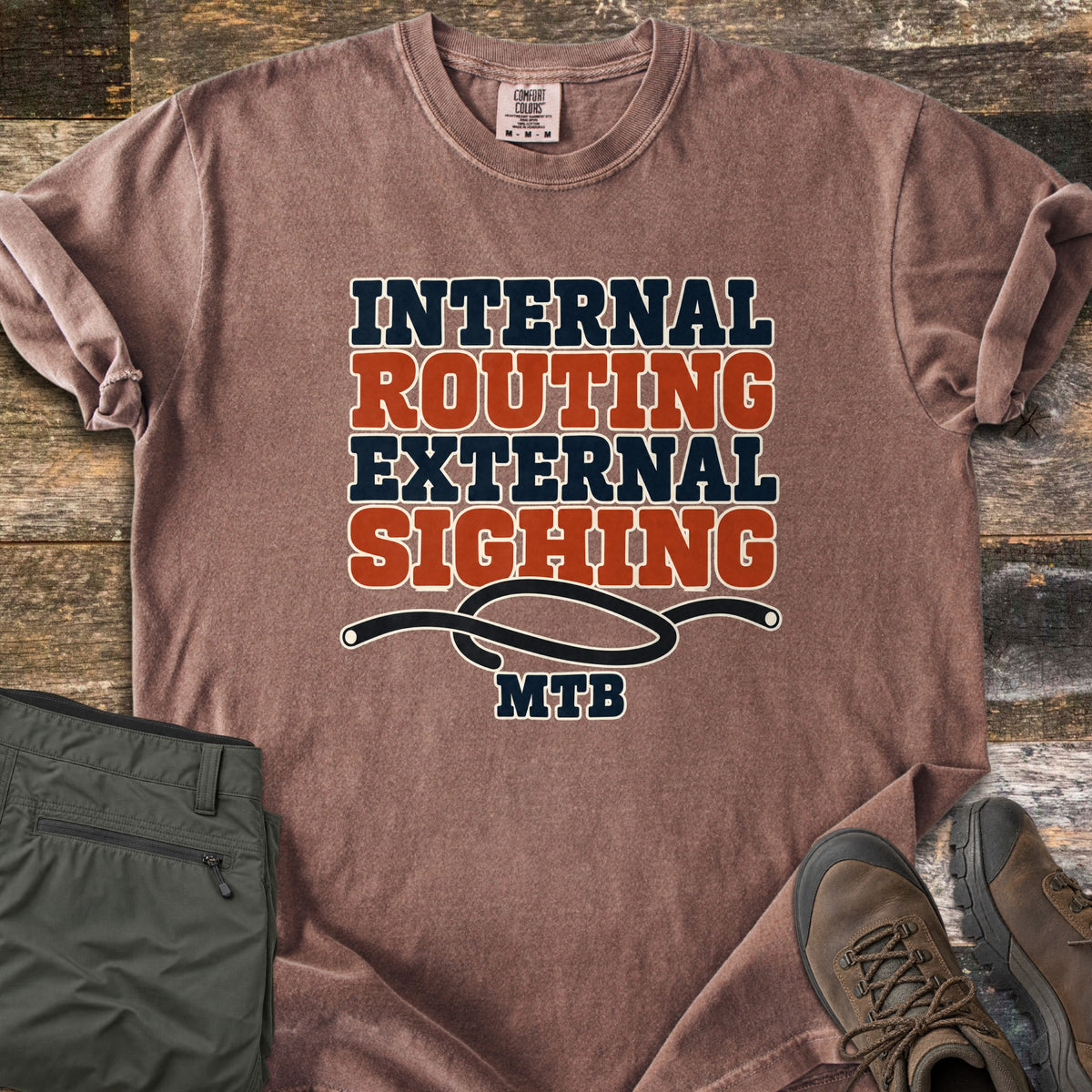 Internal Routing External Sighing Comfort Colors T-shirt in Espresso, laid flat. Block letters read Internal Routing External Sighing in navy and rust over a looped black cable and the word MTB. Sleeves rolled, on weathered wood with folded olive pants and brown hiking boots.
