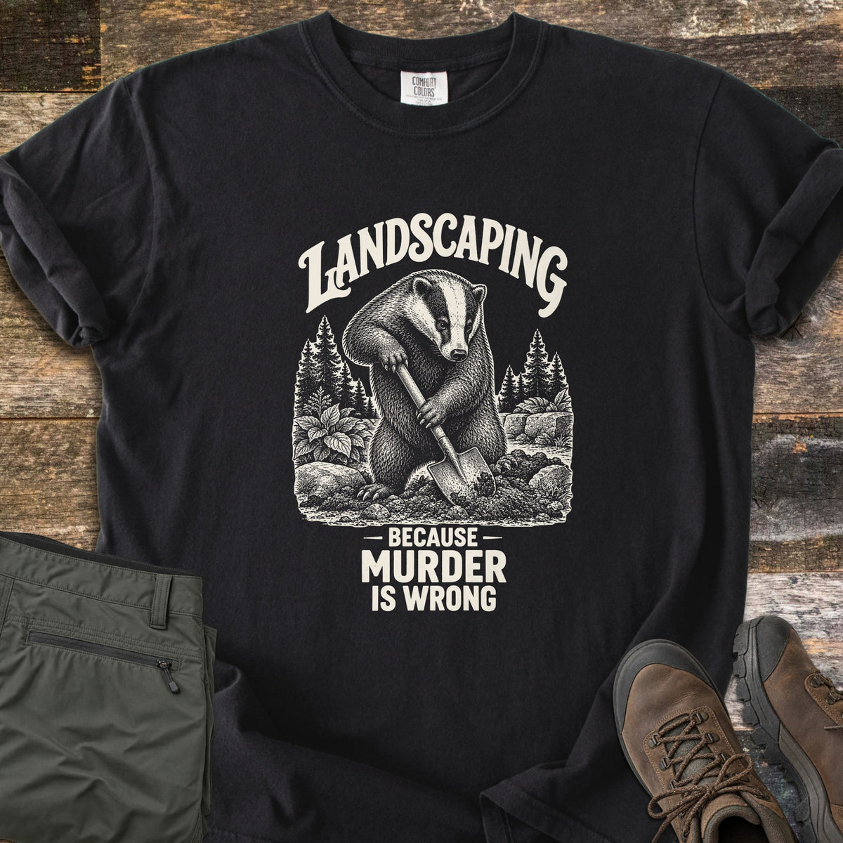 Landscaping Because Murder Is Wrong Comfort Colors T-shirt in Black, laid flat. A white engraving-style badger digs with a shovel among pine trees, plants and rocks under the arched word Landscaping, above Because Murder Is Wrong. Set on weathered wood beside olive hiking pants and brown boots.