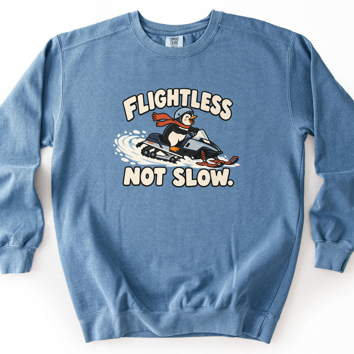 Flightless Not Slow Comfort Colors crewneck sweatshirt in Blue Jean, laid flat. A smiling cartoon penguin in a blue-gray helmet and red scarf rides a snowmobile through spraying snow, with Flightless above and Not Slow. below in cream letters. It lies on a white background.