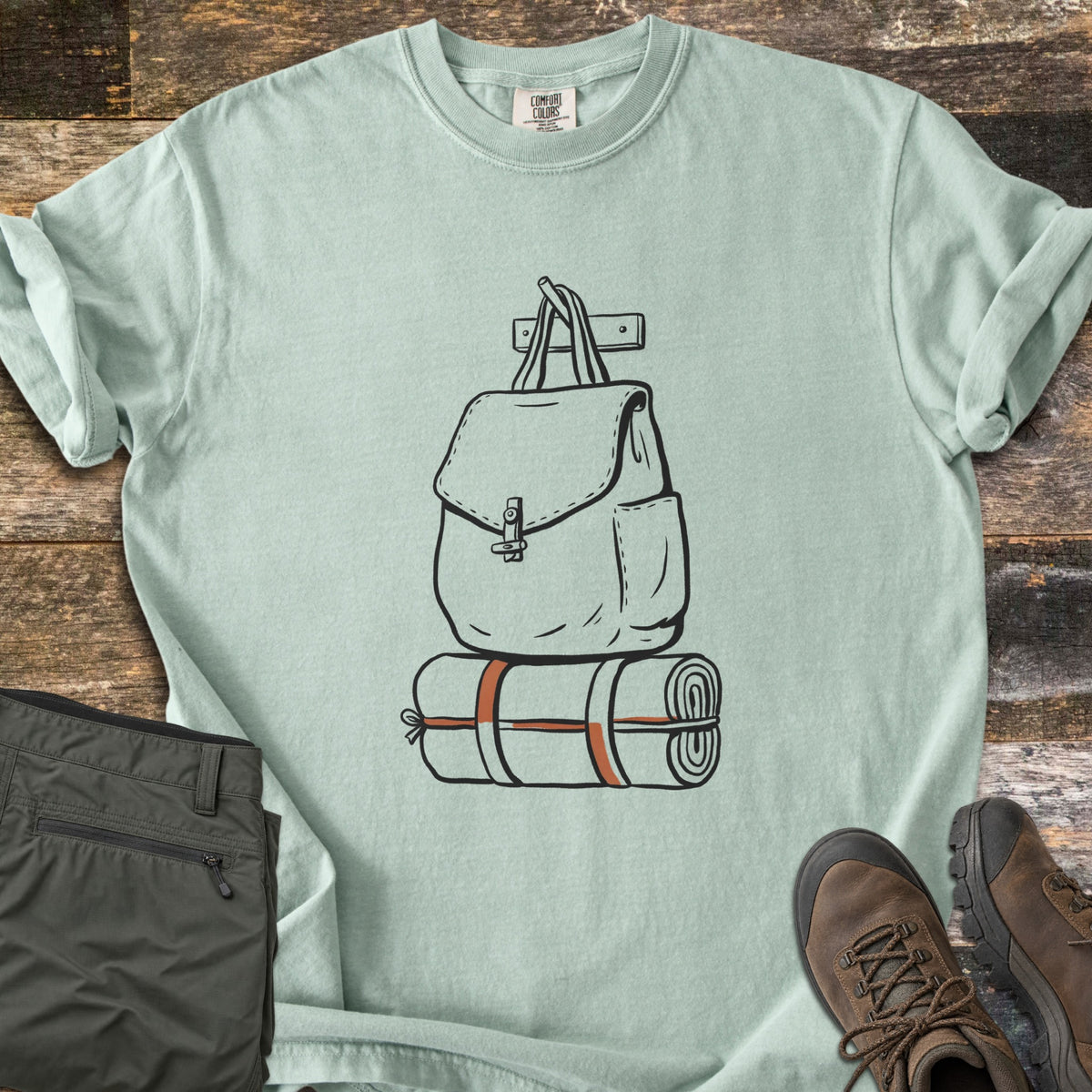 Backpack And Bedroll Comfort Colors T-shirt in Bay, laid flat. Black line art of a canvas backpack hanging from a wall peg above a bedroll tied with rust-orange straps. Olive hiking pants and brown hiking boots sit beside it on weathered wood planks.