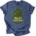 Moss Impressions Comfort Colors T-shirt in Heather Navy, laid flat. A large fingerprint made of swirling ridges of green moss, with the words Moss Impressions in cream lettering across its lower half. The shirt has rolled sleeves and lies on a white background.