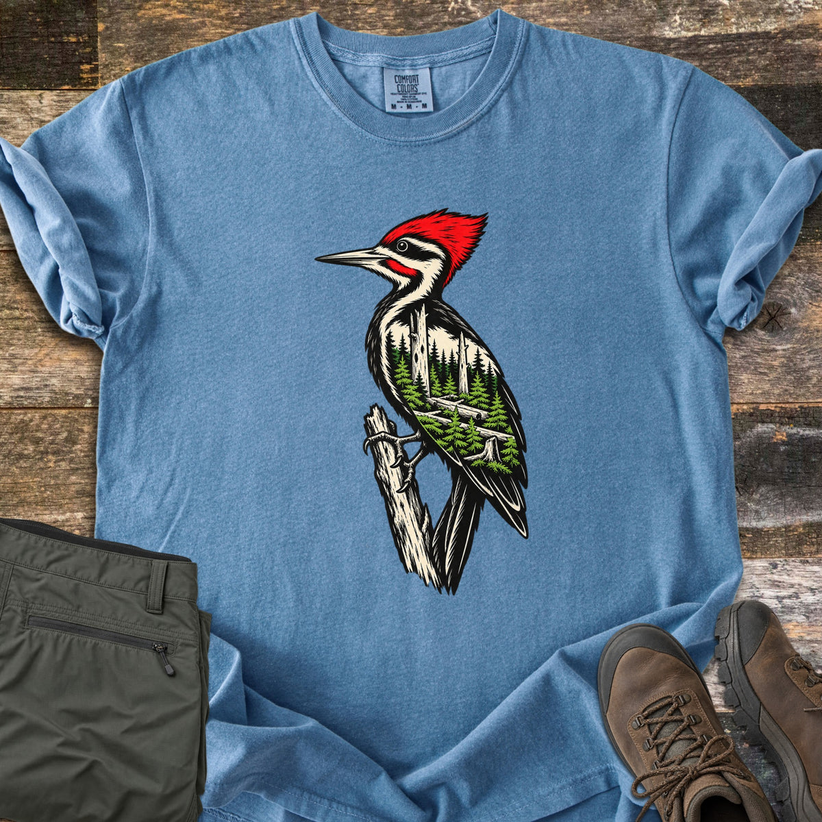 Woodpecker Forest Comfort Colors T-shirt in Blue Jean, laid flat. A pileated woodpecker with a red crest perches on a broken branch, its body filled with a pine forest scene. The shirt has rolled sleeves on weathered wood beside folded olive pants and brown hiking boots.