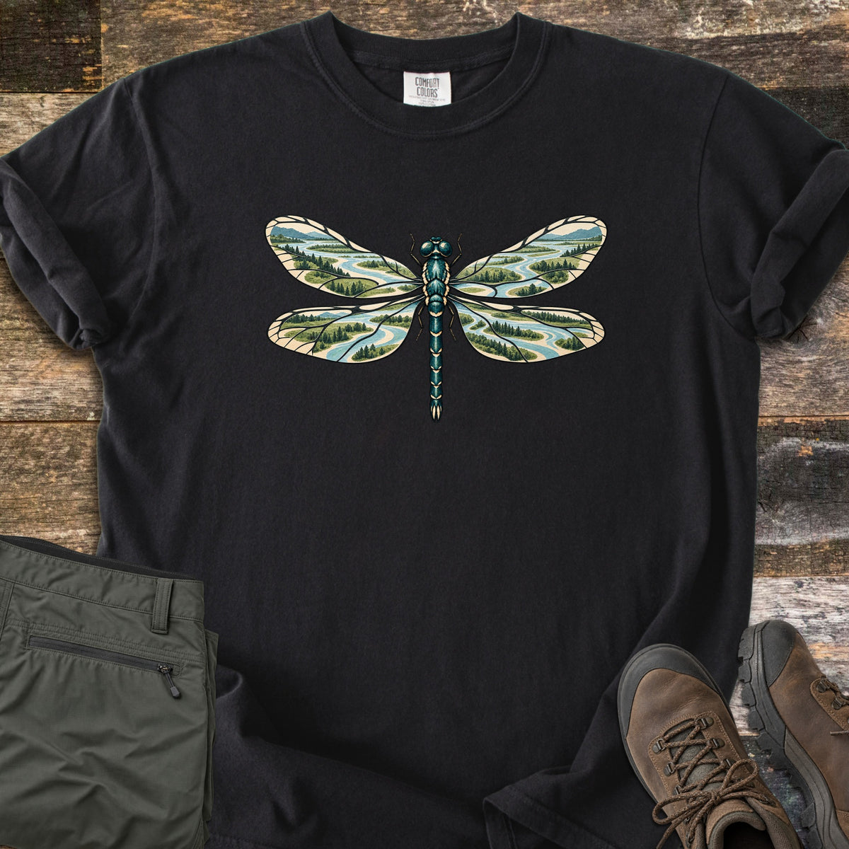 Braided River Dragonfly Comfort Colors T-shirt in Black, laid flat. A dragonfly with a teal body and wings filled with braided rivers, forests and mountains is printed on the chest, with no words. Olive cargo pants and brown hiking boots lie beside it on weathered wood.