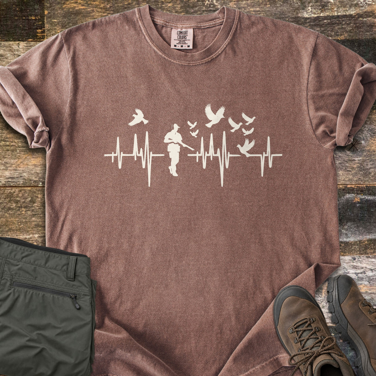 Bird Hunter Comfort Colors T-shirt in Espresso, laid flat. A cream heartbeat line crosses the chest with a hunter silhouette in a cap holding a shotgun and birds flying up from the line. Folded olive pants and brown hiking boots lie beside it on weathered wood.