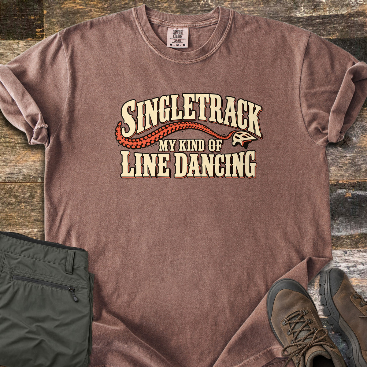 Singletrack My Kind of Line Dancing Comfort Colors T-shirt in Espresso, laid flat. Cream western-style lettering reads Singletrack, My Kind of Line Dancing, with an orange tire track curving to a bike helmet. Shown on weathered wood with folded green hiking pants and brown hiking boots.