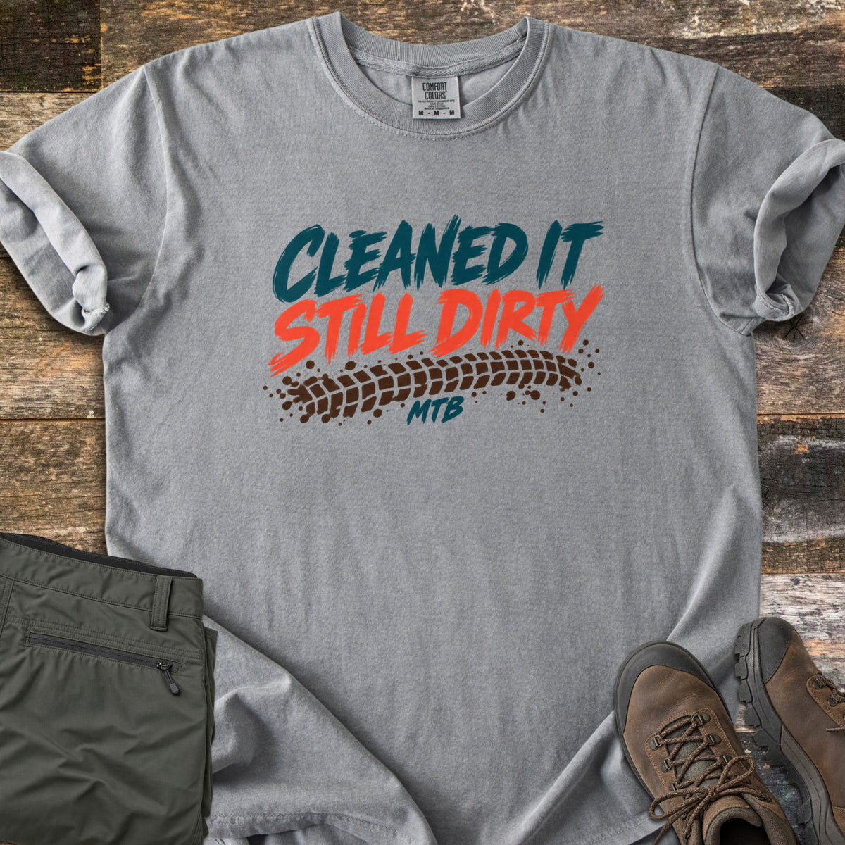 Cleaned It Still Dirty Comfort Colors T-shirt in Grey, laid flat. Brushstroke lettering reads Cleaned It in teal and Still Dirty in orange above a muddy brown tire track and the word MTB. Olive hiking pants and brown hiking boots sit beside it on weathered wood planks.