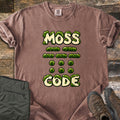 Moss Code Comfort Colors T-shirt in Espresso, laid flat. Moss-covered cream bubble letters spell MOSS, then moss-tuft dashes and dots spelling MOSS in Morse code, then CODE. It lies on weathered wood beside folded olive hiking pants and a brown hiking boot.