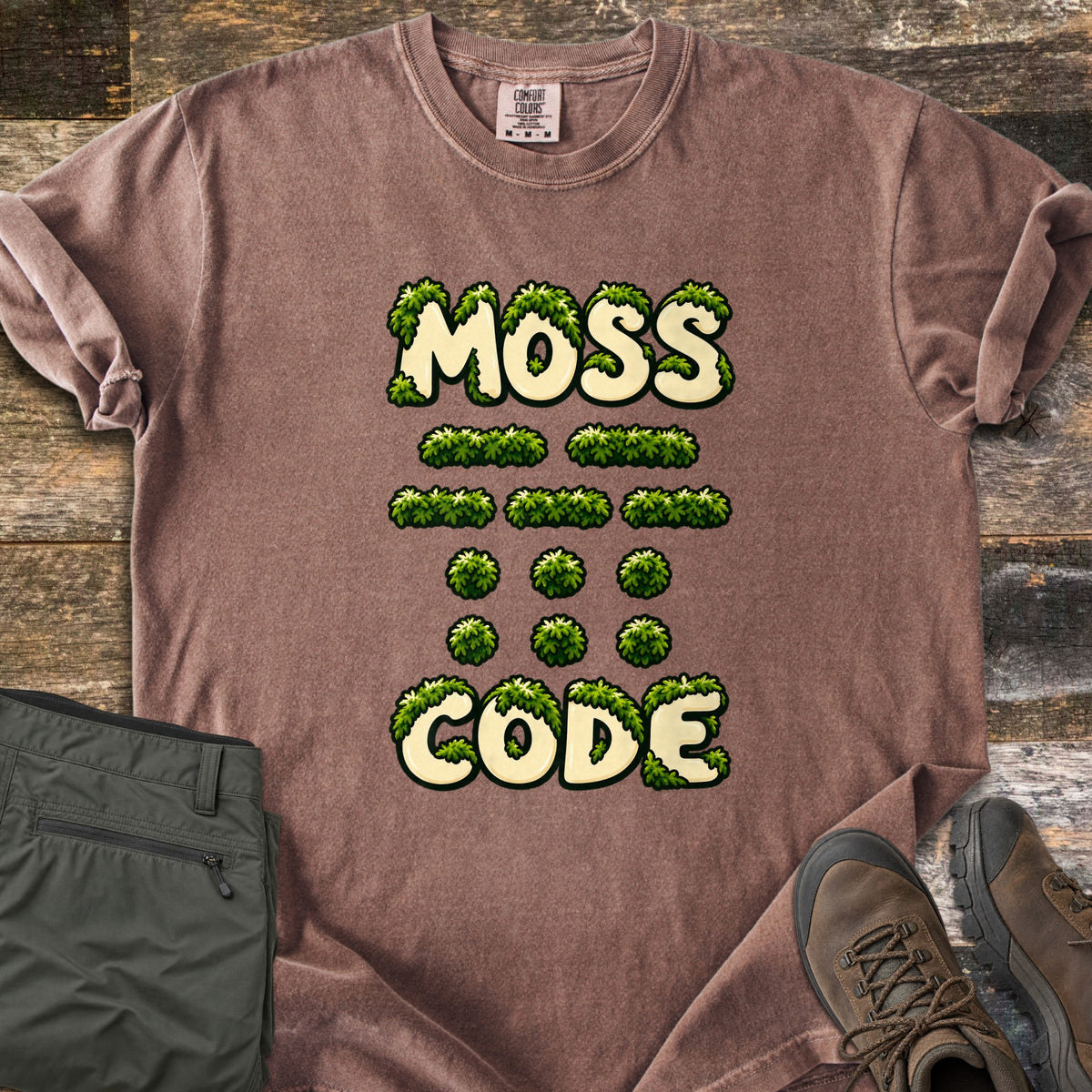 Moss Code Comfort Colors T-shirt in Espresso, laid flat. Moss-covered cream bubble letters spell MOSS, then moss-tuft dashes and dots spelling MOSS in Morse code, then CODE. It lies on weathered wood beside folded olive hiking pants and a brown hiking boot.