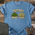 Neighbors Not Twins Comfort Colors T-shirt in Blue Jean, laid flat. Arched text Neighbors Not Twins over two illustrated rock clumps: green moss with brown stalks labeled Moss, and pale green leafy lichen labeled Lichen. Olive hiking pants and brown boots sit beside it on weathered wood.
