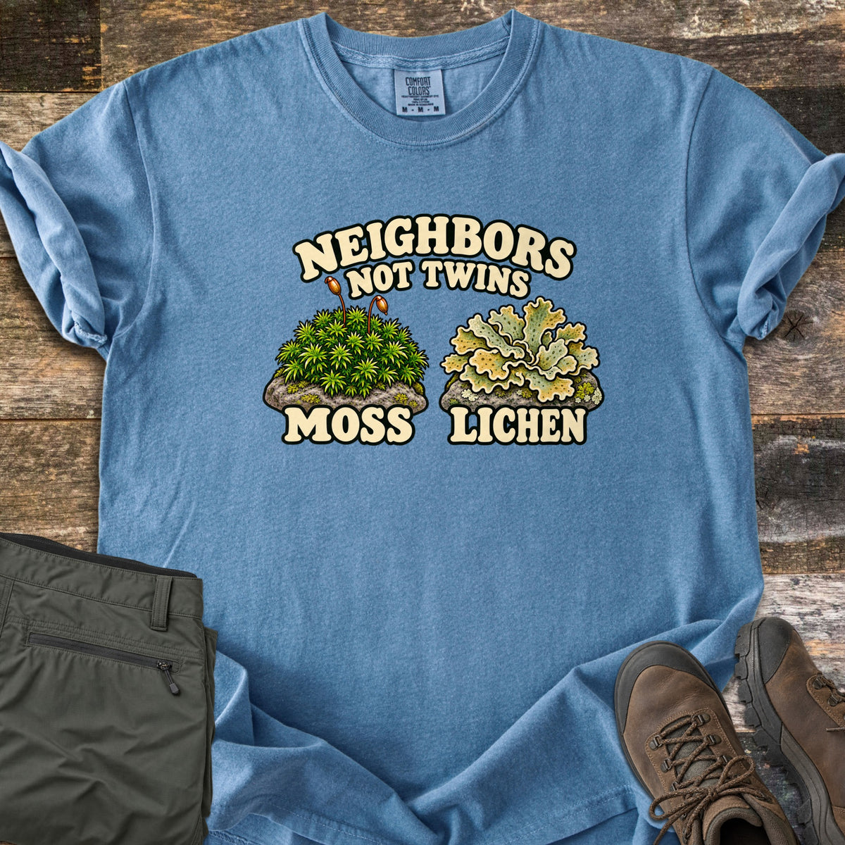 Neighbors Not Twins Comfort Colors T-shirt in Blue Jean, laid flat. Arched text Neighbors Not Twins over two illustrated rock clumps: green moss with brown stalks labeled Moss, and pale green leafy lichen labeled Lichen. Olive hiking pants and brown boots sit beside it on weathered wood.