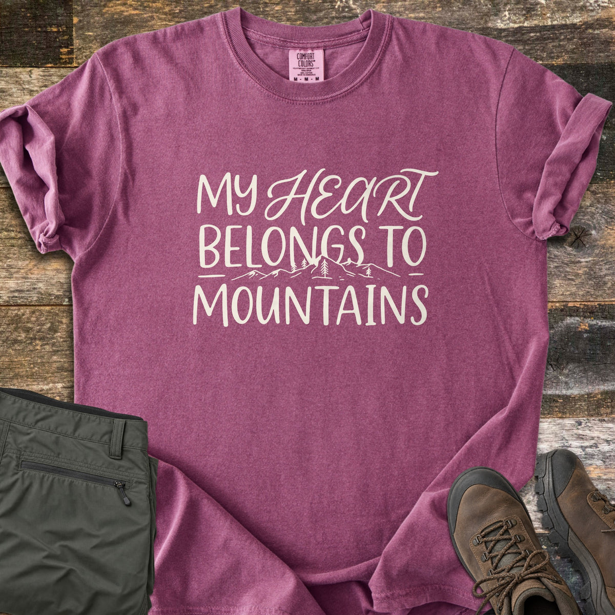 My Heart Belongs to Mountains Comfort Colors T-shirt in Berry, laid flat. Cream hand lettering reads My Heart Belongs to Mountains, with Heart in script above a line drawing of peaks and pine trees. Folded olive hiking pants and brown hiking boots sit on weathered wood.