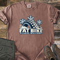 Fat Bike Flakes Welcome Comfort Colors T-shirt in Espresso, laid flat. A fat bike tire half buried in snow with three snowflakes above it, over the words FAT BIKE FLAKES WELCOME. Sleeves are rolled, on a wood floor with folded olive pants and brown hiking boots.