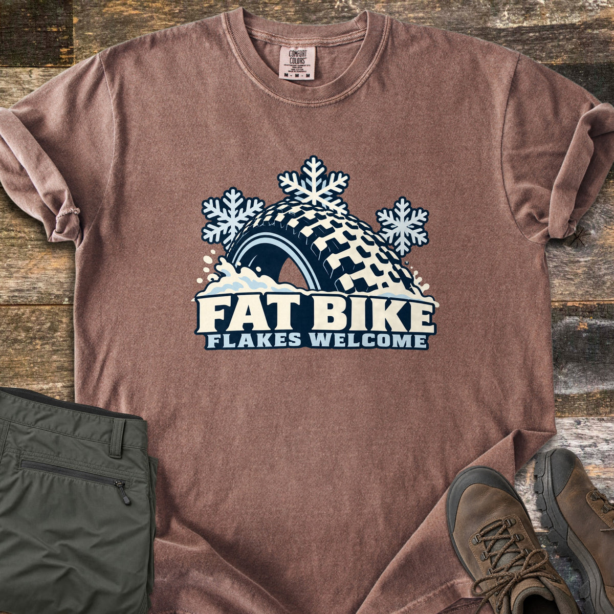 Fat Bike Flakes Welcome Comfort Colors T-shirt in Espresso, laid flat. A fat bike tire half buried in snow with three snowflakes above it, over the words FAT BIKE FLAKES WELCOME. Sleeves are rolled, on a wood floor with folded olive pants and brown hiking boots.