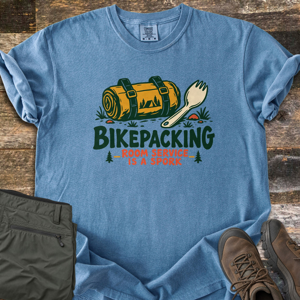 Bikepacking Room Service Is a Spork Comfort Colors T-shirt in Blue Jean, laid flat. A strapped yellow bedroll printed with a tent and pines lies beside a cream spork above the words Bikepacking Room Service Is a Spork. It rests on weathered wood with olive pants and brown hiking boots.