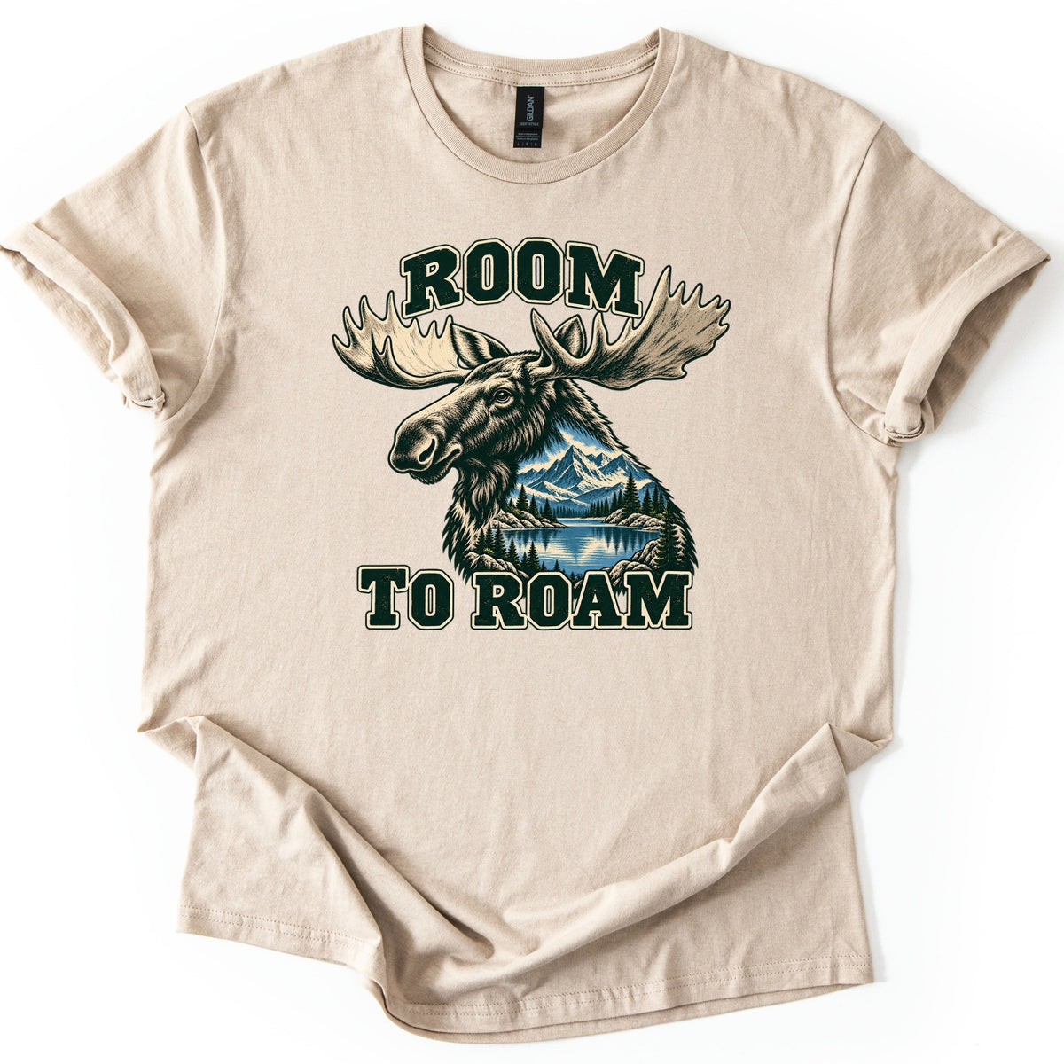 Room to Roam Comfort Colors T-shirt in Sand, laid flat. A moose head with large pale antlers, its neck filled with a scene of snowy mountains, pine trees and a blue lake, between the words ROOM and TO ROAM in dark green block letters. Sleeves rolled, on a white background.