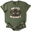 Mud Magnet Comfort Colors T-shirt in Military Green, laid flat. Bold cream letters read MUD MAGNET around a horseshoe magnet pulling brown mud splashes toward a rider on an ATV. The shirt has rolled sleeves and lies on a white background.
