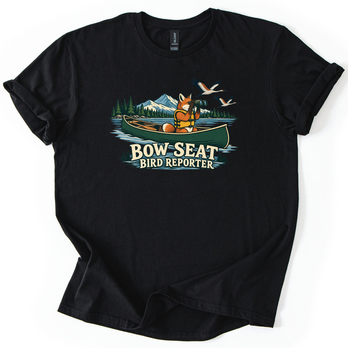 Bow Seat Bird Reporter Comfort Colors T-shirt in Black, laid flat. A fox in a life vest sits in a green canoe on a lake, looking through binoculars at flying cranes, with pines and a snowy mountain behind, above the words Bow Seat Bird Reporter. Sleeves rolled, on white.