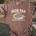 Iron Pan Cooking Co Comfort Colors T-shirt in Espresso, laid flat. Cream print of a cast iron skillet of cornbread with melting butter on a napkin, under the words Iron Pan Cooking Co. and above Seasoned Since Grandma. Folded olive pants and brown hiking boots lie on rustic wood.