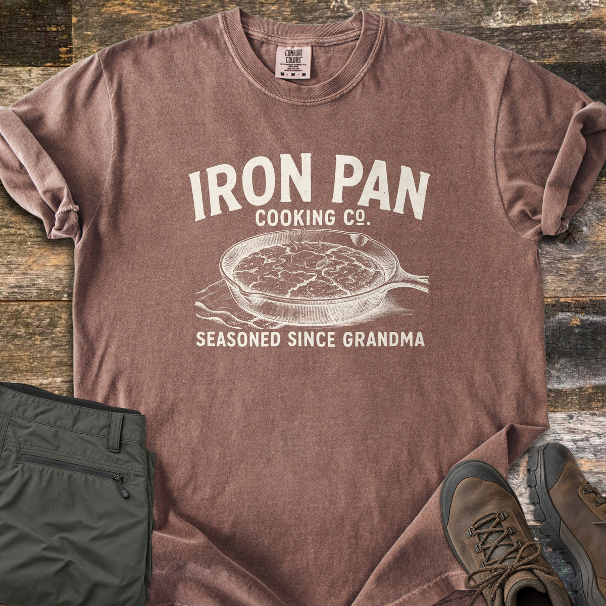 Iron Pan Cooking Co Comfort Colors T-shirt in Espresso, laid flat. Cream print of a cast iron skillet of cornbread with melting butter on a napkin, under the words Iron Pan Cooking Co. and above Seasoned Since Grandma. Folded olive pants and brown hiking boots lie on rustic wood.