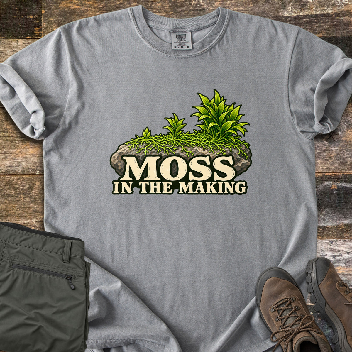 Moss in the Making Comfort Colors T-shirt in Grey, laid flat. A mossy gray rock sprouting green leafy plants sits above the words Moss in the Making. Olive hiking pants and brown hiking boots lie beside it on weathered wood planks.