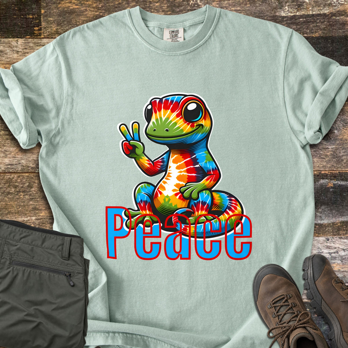 Peace Comfort Colors T-shirt in Bay, laid flat. A smiling rainbow tie-dye gecko sits flashing a peace sign above the word Peace in blue letters outlined in red. Shown on weathered wood with folded olive pants and brown hiking boots.