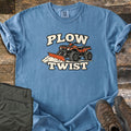 Plow Twist Comfort Colors T-shirt in Blue Jean, laid flat. An orange ATV with a snow plow blade pushes a pile of snow between the words Plow Twist in cream lettering. Folded olive cargo pants and brown hiking boots sit beside it on weathered wood planks.
