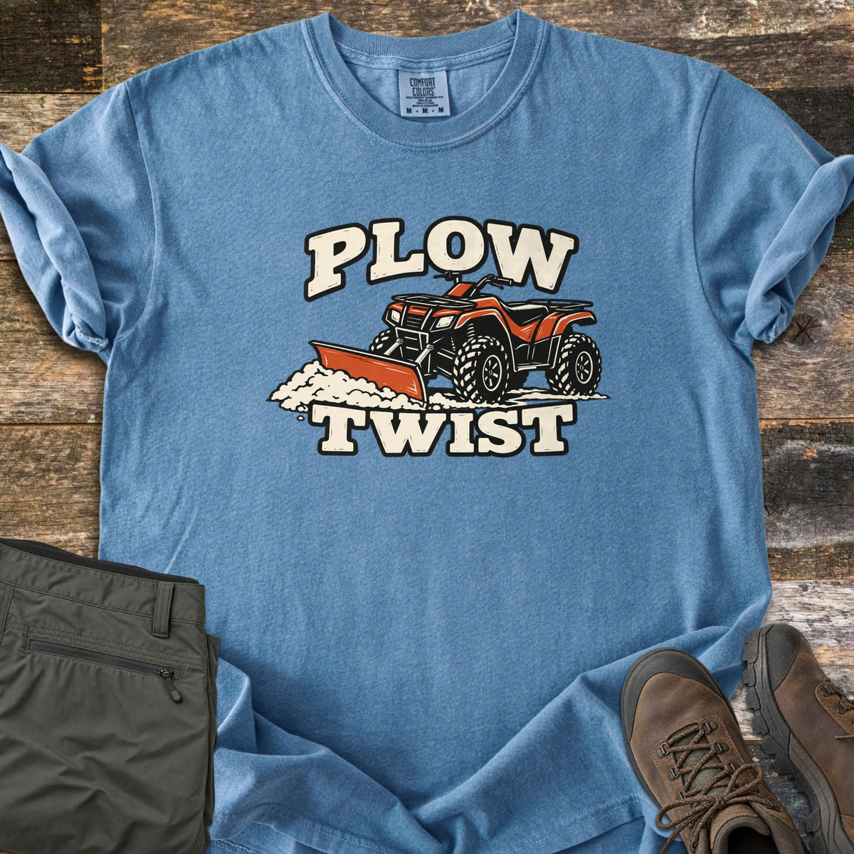 Plow Twist Comfort Colors T-shirt in Blue Jean, laid flat. An orange ATV with a snow plow blade pushes a pile of snow between the words Plow Twist in cream lettering. Folded olive cargo pants and brown hiking boots sit beside it on weathered wood planks.
