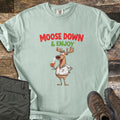 Moose Down And Enjoy Comfort Colors T-shirt in Bay, laid flat. A cartoon moose in a white shirt holds a glass of orange drink under the words Moose Down & Enjoy in red and green. Shown on rustic wood with folded olive pants and brown hiking boots.