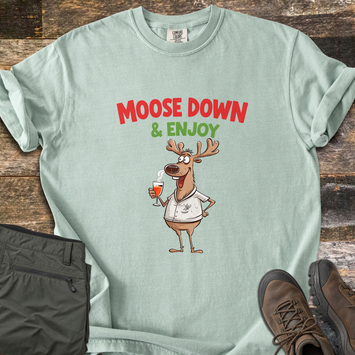 Moose Down And Enjoy Comfort Colors T-shirt in Bay, laid flat. A cartoon moose in a white shirt holds a glass of orange drink under the words Moose Down & Enjoy in red and green. Shown on rustic wood with folded olive pants and brown hiking boots.