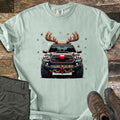 Santa Truck Comfort Colors T-shirt in Bay, laid flat. A gray pickup truck wearing reindeer antlers and a red nose, with string lights and a red bow on the bumper, among stars and snowflakes. Laid on a wood floor with olive pants and brown hiking boots.